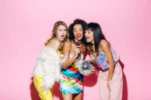 Beautiful multiethnic girls holding disco ball and champagne while singing karaoke with microphone on pink